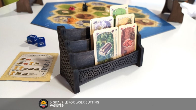 May include: A dark brown wooden card holder with four slots for holding cards. The card holder is shaped like a bench and has a decorative pattern on the front. There are five cards in the holder, each with a different image of a resource from the Settlers of Catan board game.