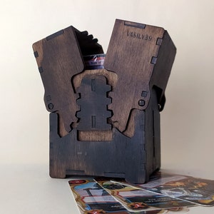 May include: A wooden box with a hinged lid, the lid is open and the box is empty. The box is stained a dark brown color and has a black finish. The box is labeled "VASILY39". There are several playing cards on the table in front of the box.