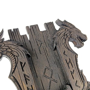 May include: A dark brown wooden wall decor featuring two dragon heads carved on either side of a central panel. The panel has carved runes. The dragons have detailed scales and open mouths.