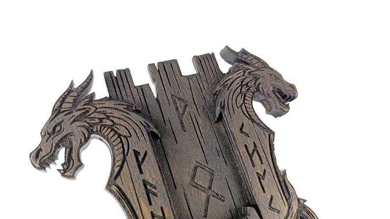 May include: A dark brown wooden wall decor featuring two dragon heads carved on either side of a central panel. The panel has carved runes. The dragons have detailed scales and open mouths.