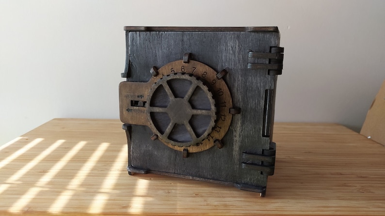 May include: A wooden box shaped like a safe with a metal dial and a handle. The dial has numbers 1 through 13 and the handle has a lever that can be moved up and down. The box is painted a dark grey colour and has a distressed look.