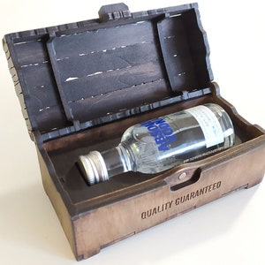 May include: A wooden box with a hinged lid, open to reveal a small bottle of Absolut Vodka. The box is stained a dark brown and has the words "Quality Guaranteed" engraved on the front.