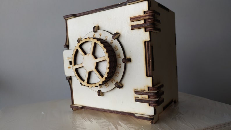 May include: A wooden toy safe with a combination lock. The safe is painted white and has a brown wooden dial with numbers 1 through 12. The safe has a small handle on the side.