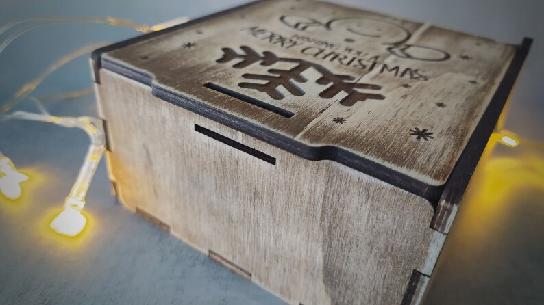 May include: A wooden box with a Christmas tree design and the text "Merry Christmas" on the top. The box is made of light brown wood and has a dark brown trim.