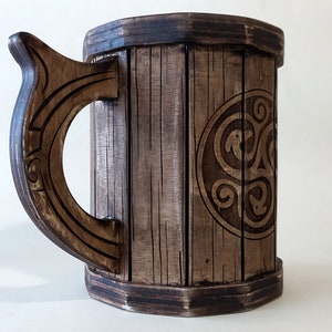 May include: A wooden beer stein with a dark brown finish. The stein has a carved handle and a Celtic knot design on the front.