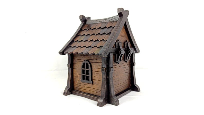 May include: A small, brown, wooden-style house model featuring a dark brown shingled roof, vertical support beams, and a single arched window on the side. The structure has a textured finish resembling wood grain and a peaked roof design.