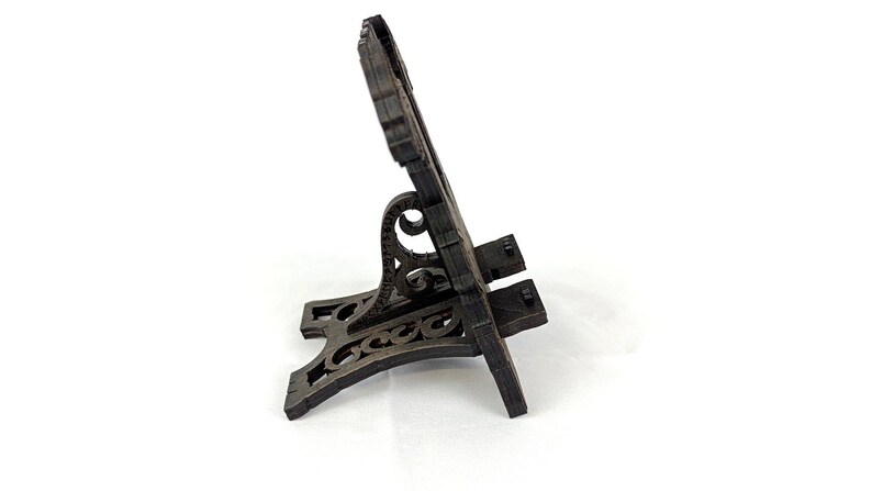 May include: A dark brown wooden phone stand with an ornate design. The stand has a decorative base and a back with a carved pattern. The stand is designed to hold a phone or small tablet.