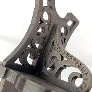 May include: Close-up of a dark brown wooden shelf bracket with intricate cut-out designs and text. The bracket has a decorative, ornate style, with a sturdy base and angled support. The wood grain is visible.