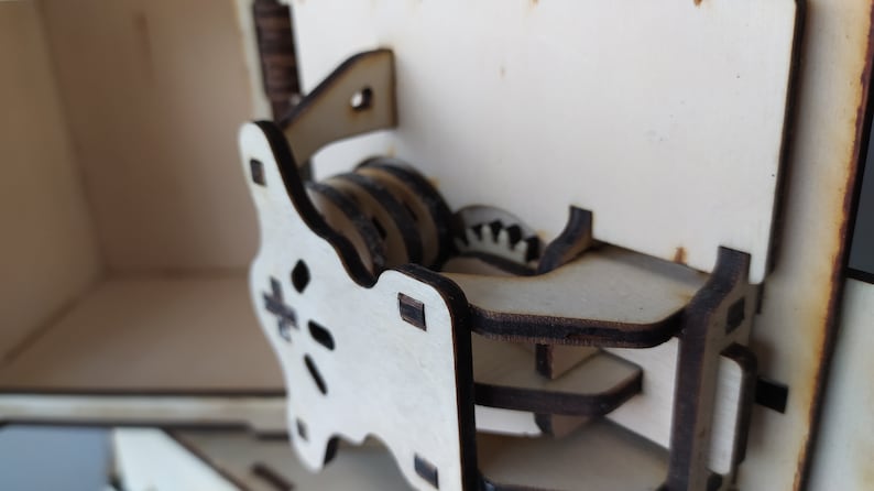 May include: Close-up of a wooden box with a complex gear mechanism. The gears are made of wood and are visible through a cutout in the box. The mechanism is designed to open the box.
