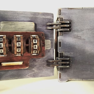 May include: A wooden box with a combination lock. The lock has three dials with numbers 1-6. The box is painted a dark gray color and has a rustic look.