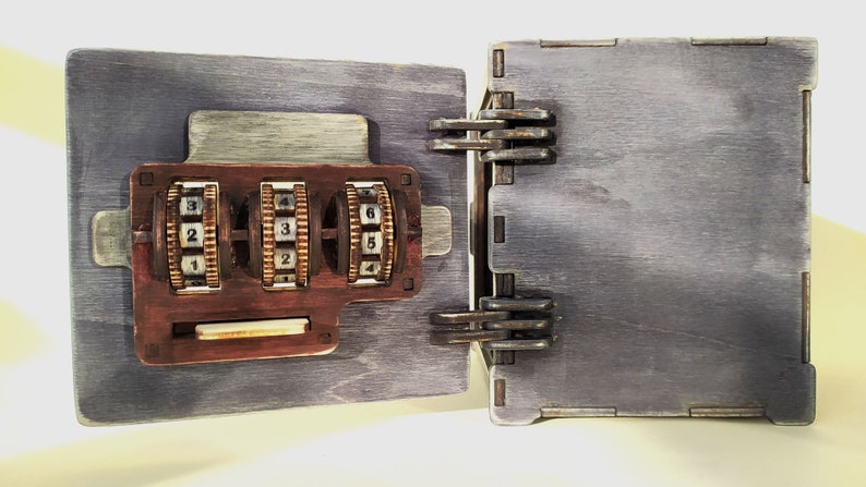 May include: A wooden box with a combination lock. The lock has three dials with numbers 1-6. The box is painted a dark gray color and has a rustic look.