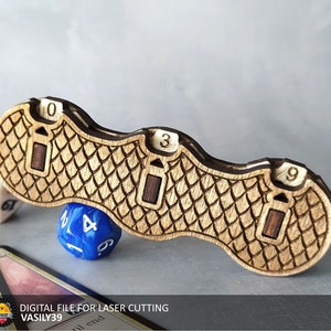 Puede incluir: Una pieza de juego de madera con un patrón de escamas y tres ranuras para dados. Las ranuras están etiquetadas con los números 0, 3 y 9. Un dado azul con el número 4 está sentado en la pieza.