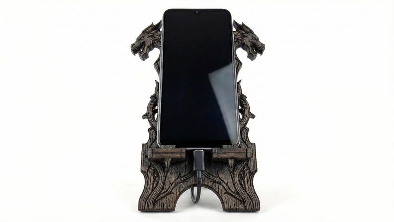 May include: A dark brown wooden phone stand with a dragon design. The stand holds a black smartphone, with a charging cable connected. The stand features two dragon heads at the top and intricate carvings.