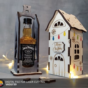 Könnte beinhalten: Ein lasergeschnittenes Holzhaus mit weißer Außenwand und braunem Dach. Das Haus hat ein Fenster, eine Tür und ein Schild mit der Aufschrift "Frohe Weihnachten". Außerdem befinden sich Lichter und Schneeflocken am Haus. Ein lasergeschnittener Holzflaschenhalter mit einer Flasche Jack Daniel's Whiskey darin.