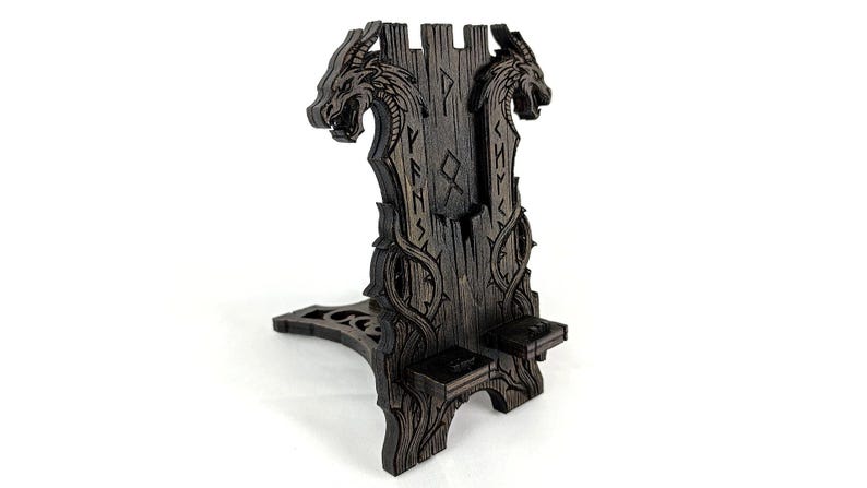 May include: A dark brown wooden phone stand with a dragon head design. The stand features carved details, including runes and a vine-like pattern. The stand is designed to hold a phone and has a unique, artistic aesthetic.