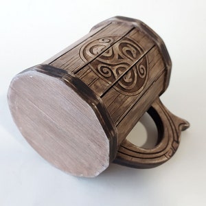 May include: A brown ceramic mug with a wooden design and a Celtic knot pattern. The mug has a handle and a flat bottom.