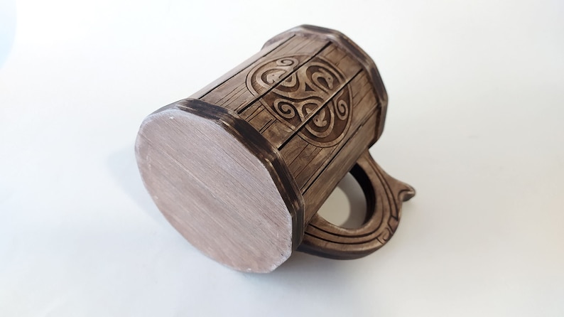 May include: A brown ceramic mug with a wooden design and a Celtic knot pattern. The mug has a handle and a flat bottom.