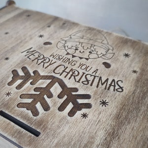 May include: A wooden box with a light brown stain. The lid has a design of a snowflake, an elf, and the text "Wishing you a Merry Christmas".