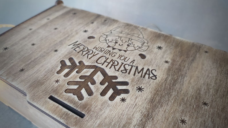 May include: A wooden box with a light brown stain. The lid has a design of a snowflake, an elf, and the text "Wishing you a Merry Christmas".