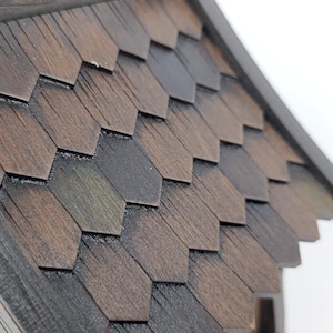 May include: A wooden birdhouse roof featuring dark brown, hexagonal shingles with a visible wood grain texture. The roof is angled, showing multiple rows of overlapping, geometric pieces that create a layered appearance on the structure.