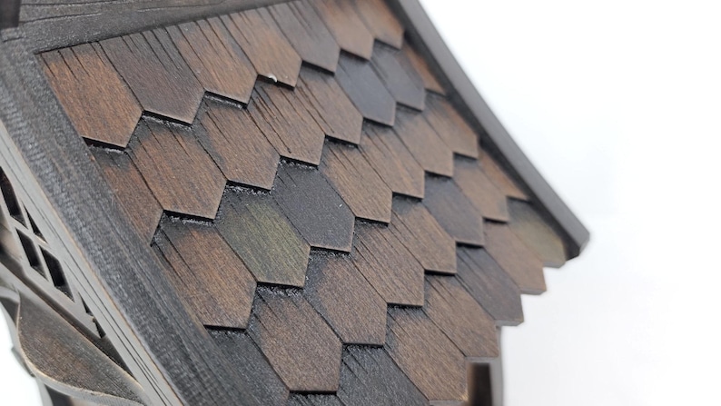 May include: A wooden birdhouse roof featuring dark brown, hexagonal shingles with a visible wood grain texture. The roof is angled, showing multiple rows of overlapping, geometric pieces that create a layered appearance on the structure.
