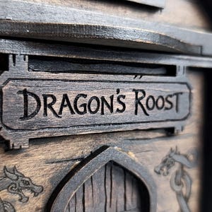 May include: A dark brown wooden structure featuring a rectangular sign with the engraved text DRAGON'S ROOST. Below the sign, a pointed archway is flanked by two engraved dragon illustrations on the wood grain surface of the object.