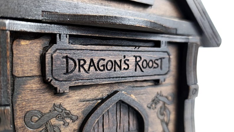 May include: A dark brown wooden structure featuring a rectangular sign with the engraved text DRAGON'S ROOST. Below the sign, a pointed archway is flanked by two engraved dragon illustrations on the wood grain surface of the object.