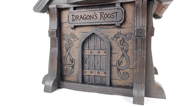 May include: A dark brown wooden structure featuring a central arched door with a keyhole, flanked by two engraved dragon motifs. Above the door, a rectangular sign is clearly inscribed with the text DRAGON'S ROOST in dark lettering.