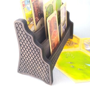 May include: A wooden card holder with a textured, dark brown finish. The holder has four slots for cards and a decorative design on the front. The holder is sitting on a table with several game tiles and dice.
