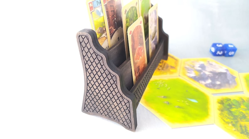 May include: A wooden card holder with a textured, dark brown finish. The holder has four slots for cards and a decorative design on the front. The holder is sitting on a table with several game tiles and dice.