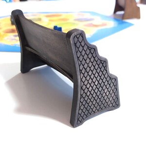 May include: A dark brown wooden game piece shaped like a bench with a decorative, textured back. The bench is designed to hold game pieces or cards.