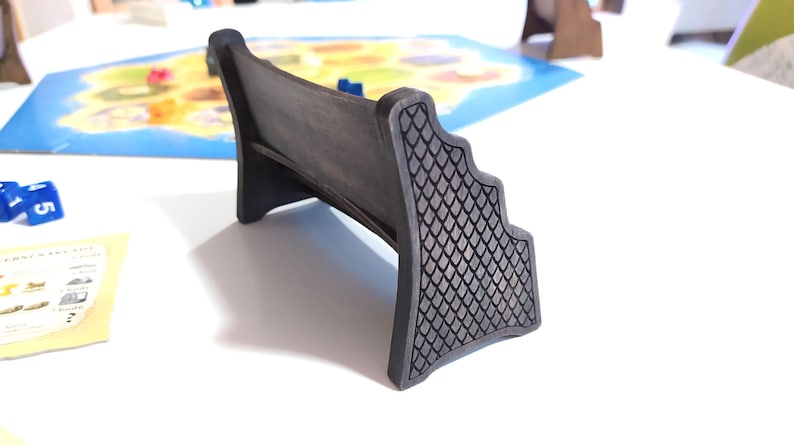 May include: A dark brown wooden game piece shaped like a bench with a decorative, textured back. The bench is designed to hold game pieces or cards.