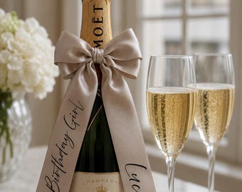 May include: A bottle of Moët & Chandon Impérial Brut champagne adorned with a beige satin ribbon tied in a bow. The ribbon features the black printed text 30th Birthday Girl and the name Lucie. Two tall champagne flutes filled with sparkling wine.