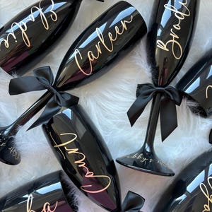 May include: A collection of glossy black champagne flutes, each featuring a personalised name in gold script lettering. Every flute is adorned with a black satin ribbon tied around the stem. The names include Elodie, Carleen, Brodie, Court, and Jacha.