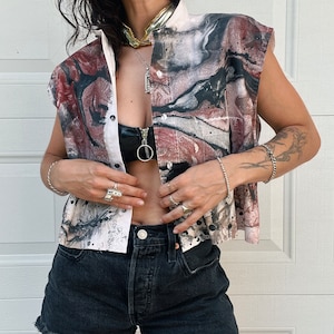 May include: A sleeveless shirt with a white base and abstract red, black, and gray marbled pattern. The shirt has a button-up collar and a single button closure. The model is wearing black denim shorts.