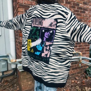 May include: A black and white zebra-striped sweater with a graphic of The Rolling Stones band members. The graphic includes the text "THE ROLLING STONES" and "Exile on Main St." The sweater has a black trim.
