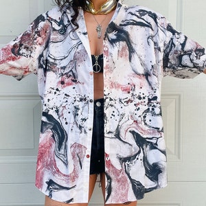 May include: White button-down shirt with an abstract pattern in black, red, and gray. The shirt has short sleeves and a classic collar. The design resembles a marble or watercolor effect, with splatters and swirls of color.