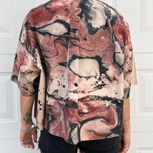 May include: Short-sleeved button-up shirt with an abstract pattern in shades of beige, burgundy, and black. The shirt has a collared neckline and is paired with black denim shorts. The overall style is casual and artistic.