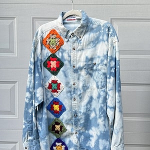 Puede incluir: Una camisa vaquera azul claro con un patrón de tie-dye y cuadrados de ganchillo de colores cosidos en la parte delantera.