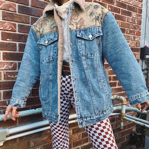 May include: A blue denim jacket with a floral tapestry design on the shoulders and a sherpa lining. The jacket is worn over a white turtleneck sweater and checkered pants.