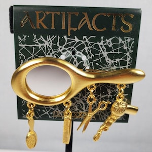 May include: Gold-toned brooch shaped like a hand mirror with dangling charms including a pair of scissors, a hairbrush, a comb, and a hairdryer. The brooch is on a black background with a green card that says "ARTIFACTS".