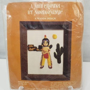 May include: A mini crewel embroidery kit featuring an Indian boy in traditional clothing, standing in front of a cactus and a pile of wood. The kit includes 100% cotton fabric, Persian yarn, a needle, a stitch chart, and simple instructions. The kit is titled "A Mini Crewel - By Needle Magic" and "A Wanda Design". The kit number is 223.