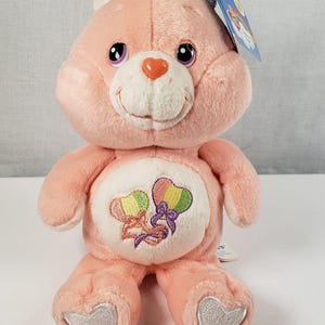 Könnte beinhalten: Rosafarbener Care Bear Plüschtier mit lila Augen und weißem Bauch. Der Bär trägt eine rosa Schleife und hat einen herzförmigen Aufnäher auf seiner Brust mit zwei Regenbogenballons.