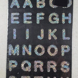 May include: A sheet of 35 iron-on sequin letters in the alphabet, A through Z, on a black background. The letters are 1 inch (2.5 cm) each. The sheet is packaged with a black t-shirt with the word "HOPE" in white sequins.