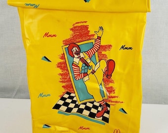 Może przedstawiać: Żółta torba na lunch z winylu z postacią Ronalda McDonalda, maskotki McDonald's, z nadrukiem słów "Mmm". Torba ma składany górny brzeg i logo McDonald's.