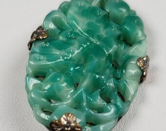 May include: A green carved jade brooch with silver accents. The brooch is oval shaped and features a floral design.