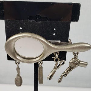 May include: Silver-toned charm bracelet featuring a hand mirror, hairbrush, comb, scissors, and a hairdryer. The charms are attached to a silver-toned chain.