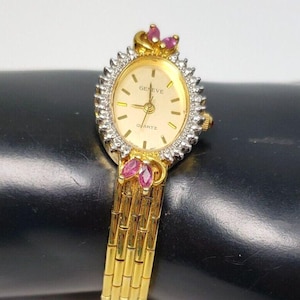 Puede incluir: Un reloj de pulsera dorado con una esfera blanca y una banda dorada. La esfera del reloj tiene un bisel de diamantes y las palabras "Geneve Quartz" impresas en él. El reloj tiene piedras preciosas rosas en la parte superior e inferior de la banda.