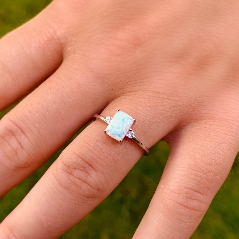 May include: A silver ring featuring a rectangular, iridescent opal center stone, flanked by two smaller, round, clear stones. The ring is set on a slender silver band, creating a delicate and elegant design. The ring is shown on a finger.