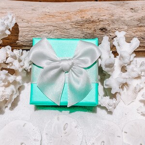 May include: A small, square, turquoise gift box with a white satin bow. The box is surrounded by white coral, sand dollars, and a piece of driftwood, creating a beachy theme. The bow is tied neatly on top of the box.
